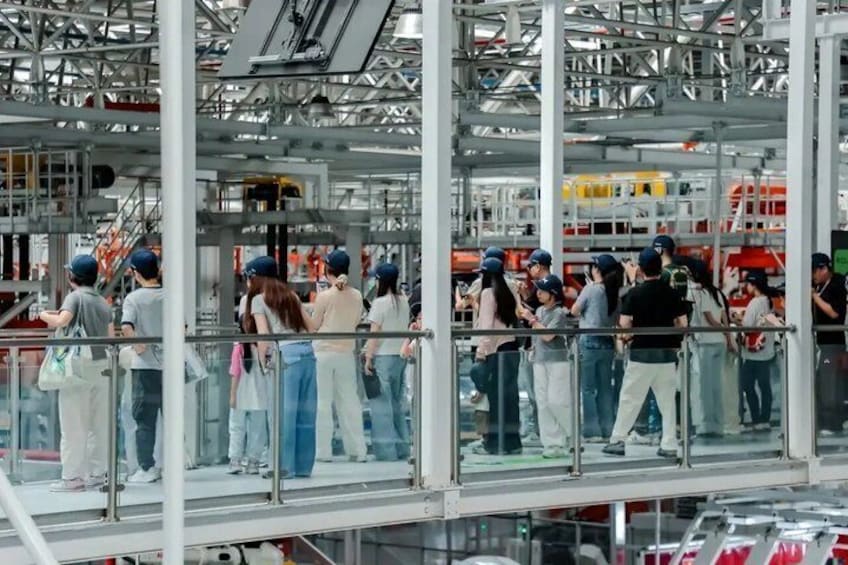 A group of people standing on a glass-walled balcony in an industrial setting with metal structures and machinery in the background. 3