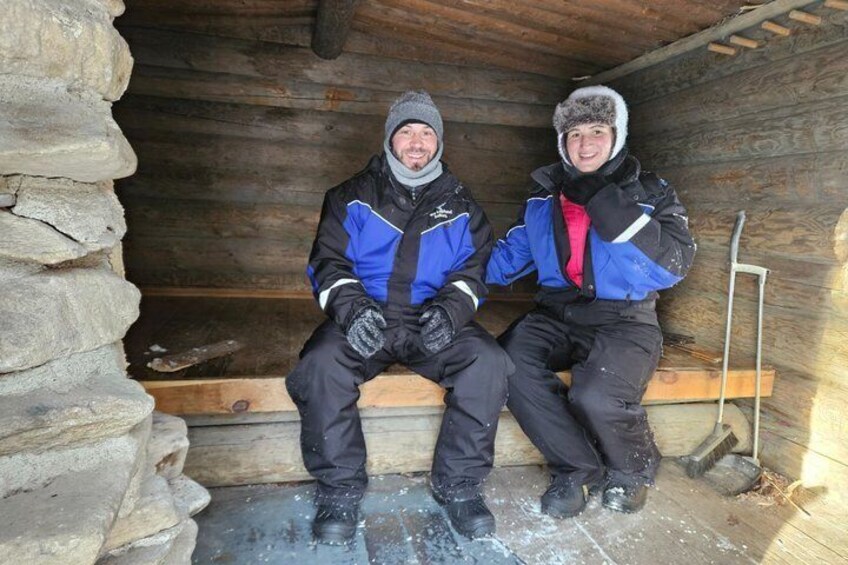 Two people sitting on a bench inside a wooden cabin with a stone wall on one side. 4