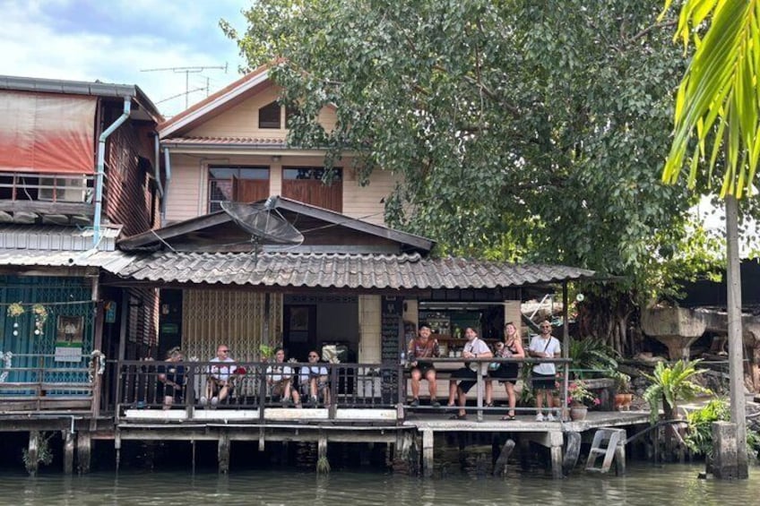 A riverside restaurant with a wooden deck and patrons seated outside. 2