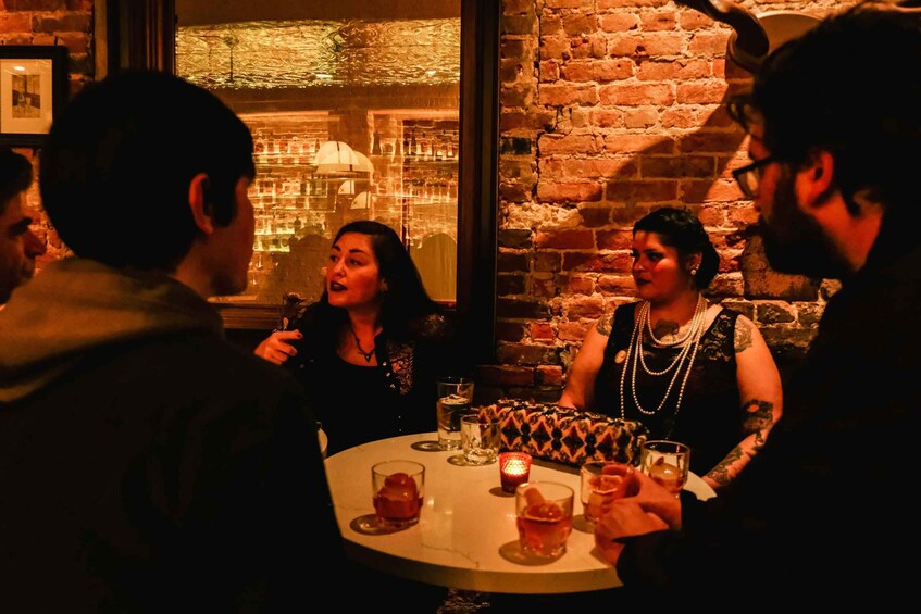 A group of people seated around a table with drinks, in a dimly lit room with brick walls and a window. 1