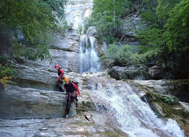A waterfall in a forested area with two people climbing a rocky path. 1