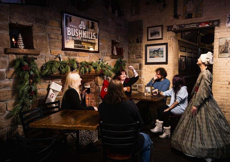 A group of people sitting at a wooden table in a rustic pub with a sign reading 'BUSHMILLS.' 2