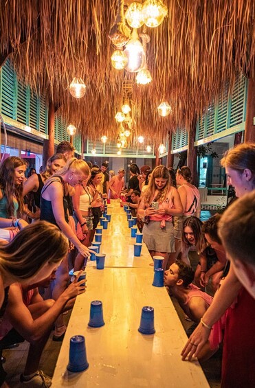 A group of people playing beer pong at a tiki bar with thatched roofs and hanging lights. 4