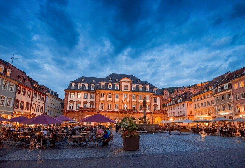 Ett historiskt torg med en framstående byggnad, uteservering och en fontän. 1