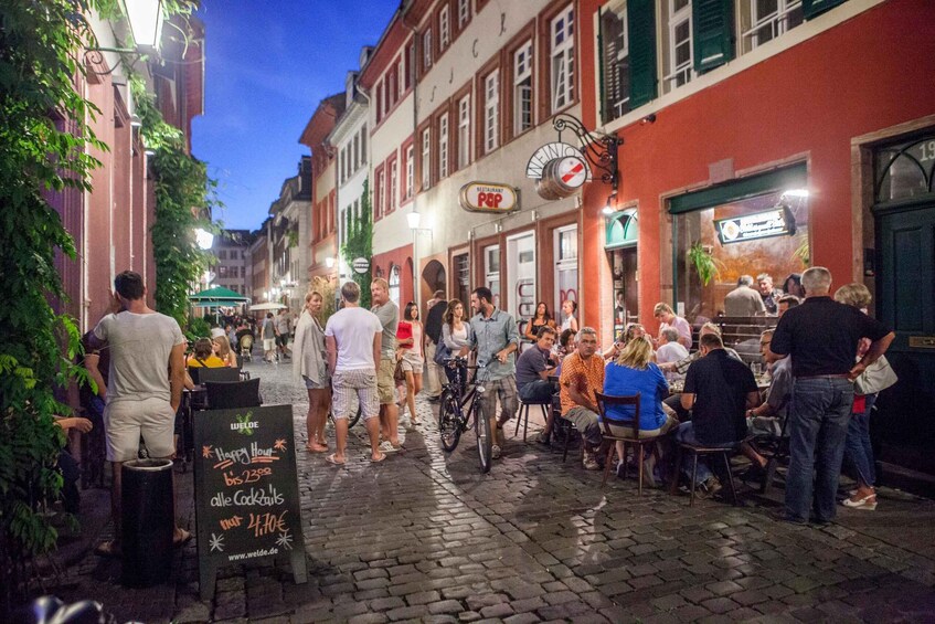 En kullerstensgata kantad av byggnader, en folkmassa och en skylt som annonserar cocktails för 4,70 euro. 2