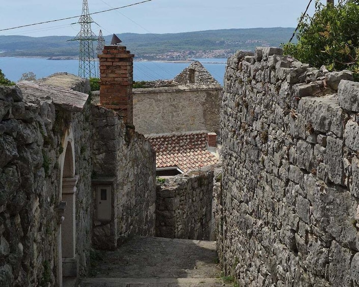 A narrow stone pathway between old stone walls, leading to a building with a tiled roof. 1