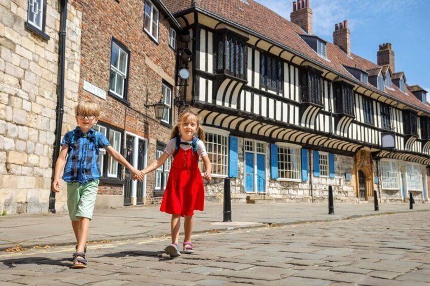 Two children holding hands walking on a cobblestone street, with historic buildings featuring timber framing and blue shutters in the background. 5