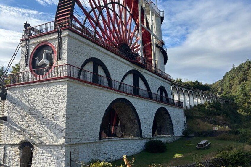 A historic water wheel with a clock tower, set against a hilly landscape. 1