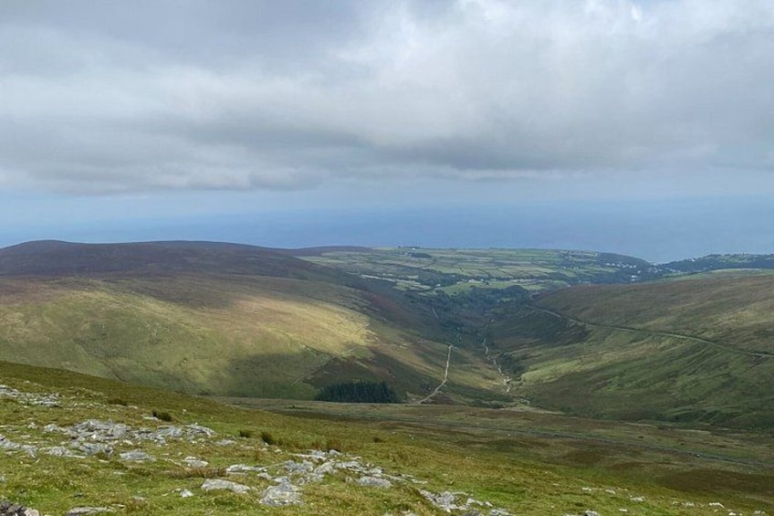 Un paysage montagneux avec une vallée, des pentes herbeuses et une vue lointaine sur la mer sous un ciel nuageux. 4