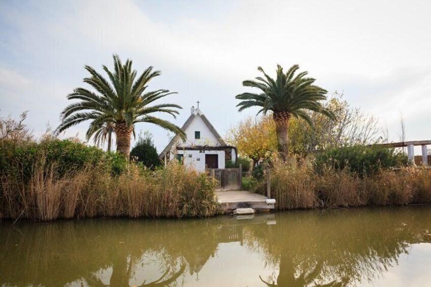 A white church with a pointed roof, surrounded by palm trees and tall grasses, with a calm body of water in the foreground. 5