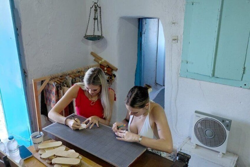 Two women working at a wooden table with tools, in a room with a fan and a blue door. 2