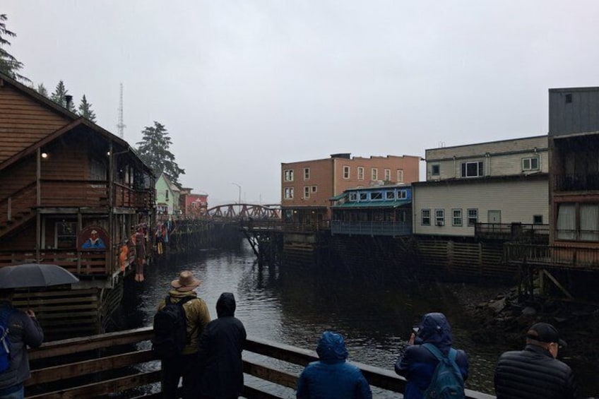 A river with wooden buildings on the left and a bridge in the background. 1