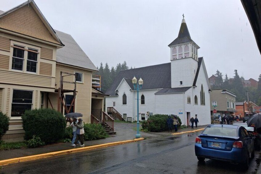 A church with a steeple, a blue car, and a person with an umbrella on a wet street. 4
