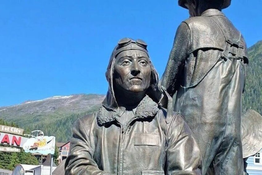 A statue of a man in a leather jacket and a woman in a dress, with a mountainous background and a sign that reads '1st Class.' 2