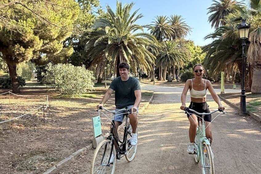 Two people riding bicycles on a path surrounded by trees and palm trees. 4