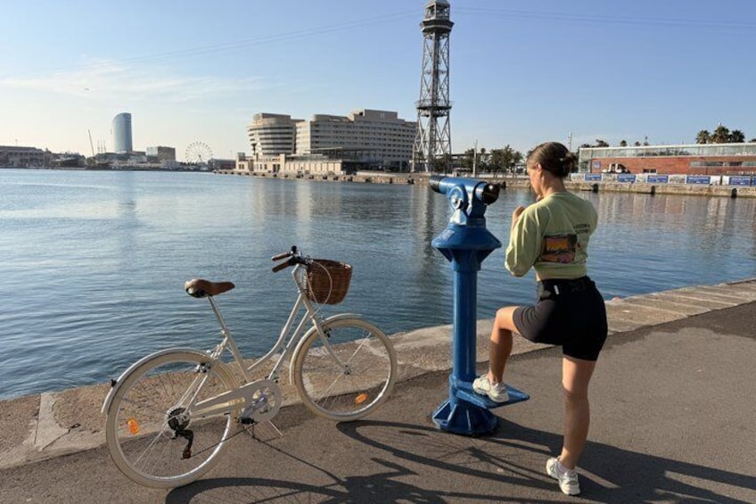 A person standing by a waterfront, looking through a telescope, with a bicycle parked nearby. 1