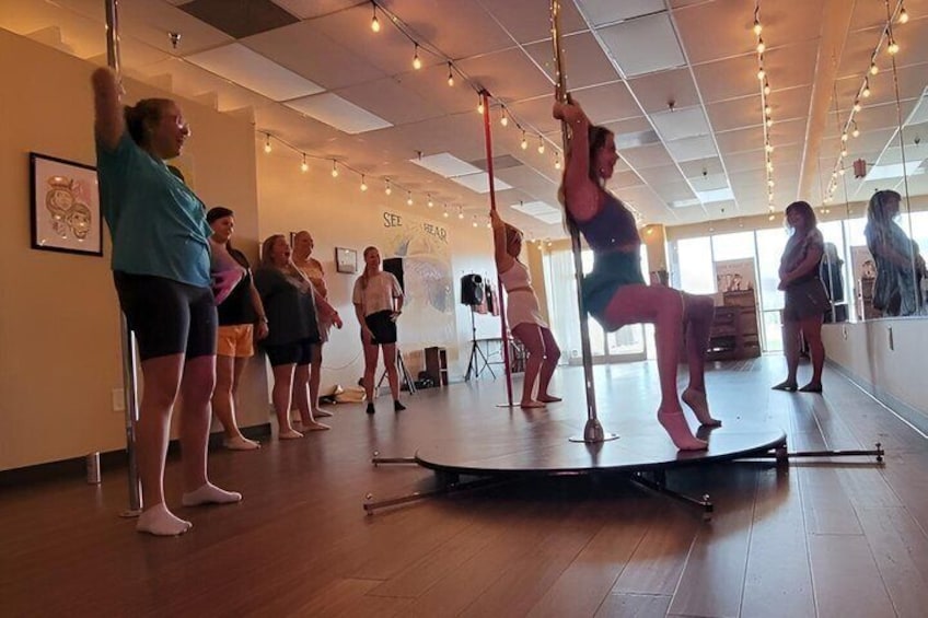 A dance studio with a pole in the center, people practicing pole dancing, and a mirror on the wall. 5