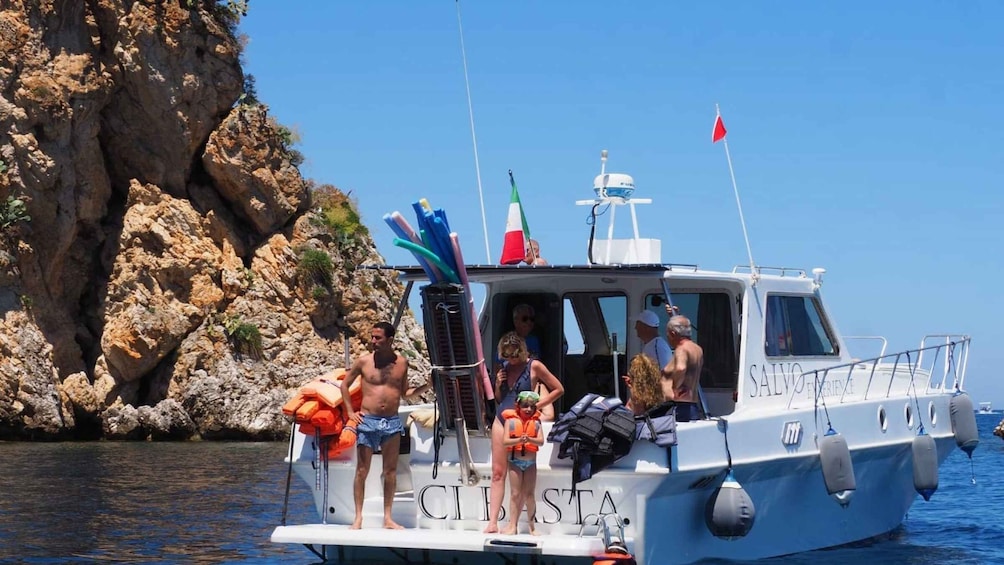 A boat named SALVO with people on board, a rocky coastline, and a clear blue sky. 1