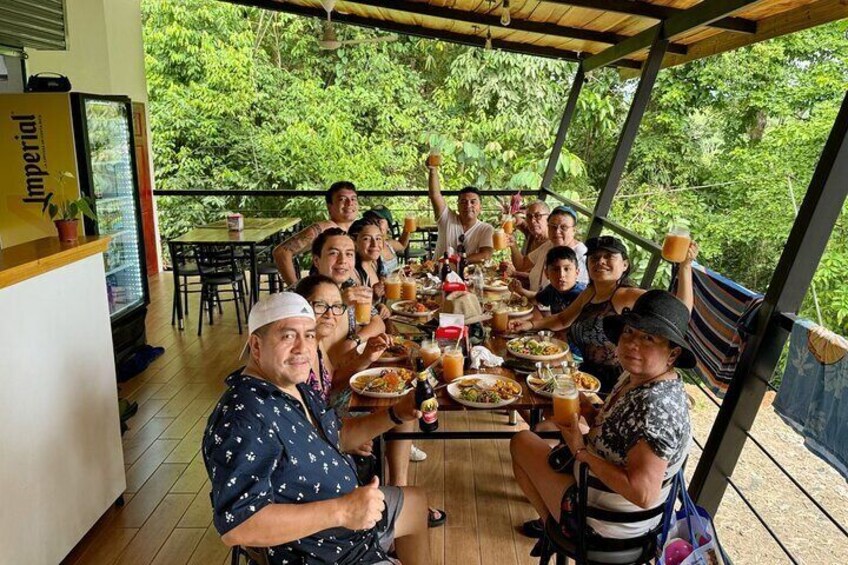 A group of people dining outdoors at a wooden table with a view of lush greenery. 2