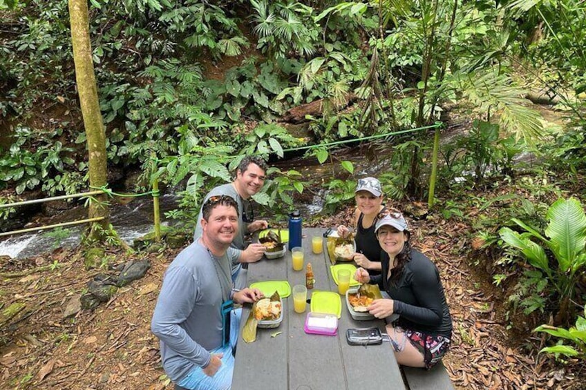 Four people sitting at a table outdoors with food and drinks, surrounded by dense greenery and a small stream. 3