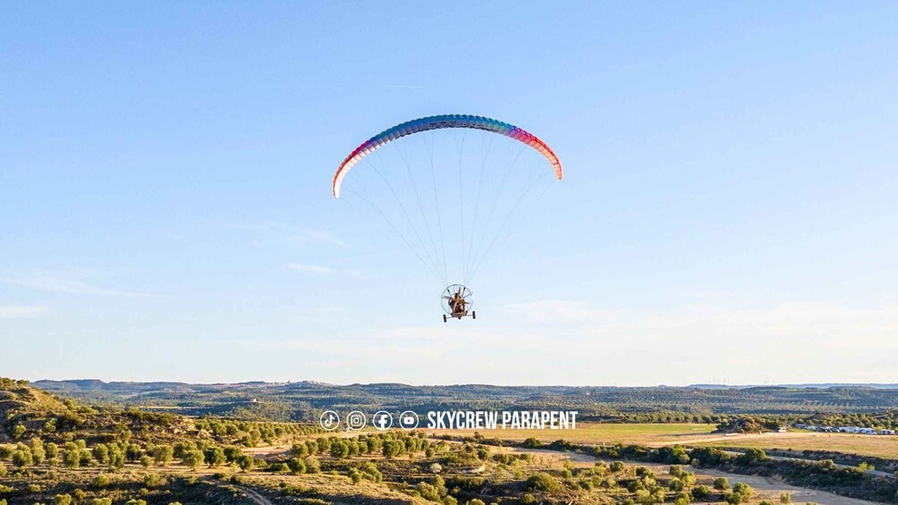 A paraglider in mid-flight over a rural landscape with rolling hills and sparse vegetation. 3