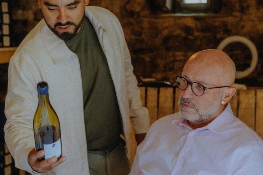 A person holding a bottle of wine, another person seated, and a rustic interior with wooden elements. 1