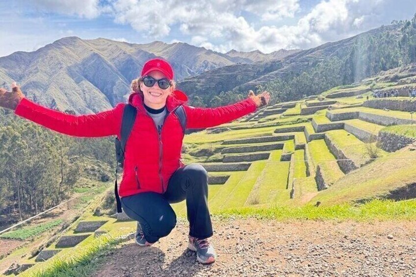 A person in a red jacket and black pants squatting on a dirt path with a mountainous landscape in the background. 5