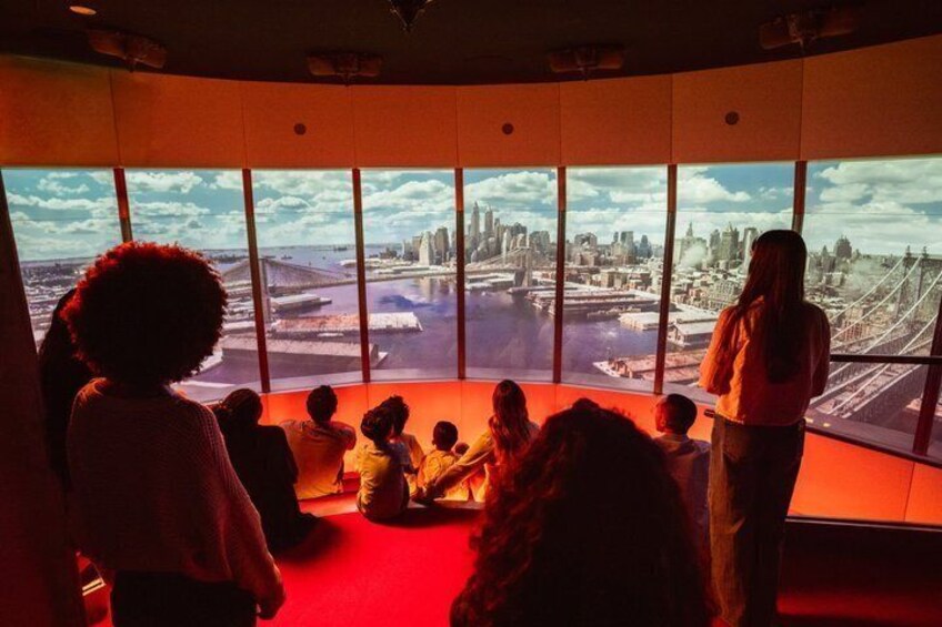A group of people sitting and standing in a room with large windows, looking at a projected image of a cityscape. 3