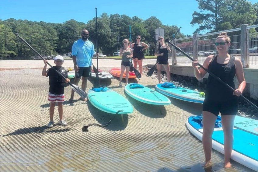 People standing on a dock with paddleboards, a calm body of water, and trees in the background. 2