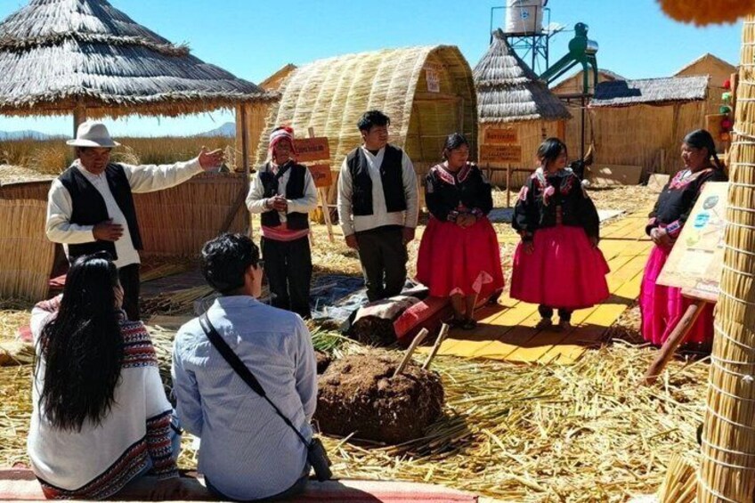 A traditional dance performance in a rural setting with straw-covered ground and thatched-roof structures. 3
