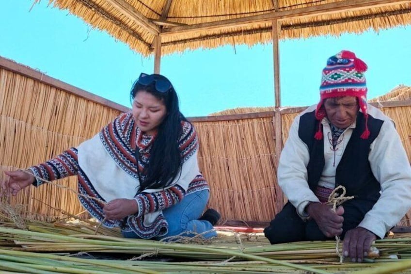 Two people sitting outdoors, one holding a rope, with a thatched roof structure in the background. 5
