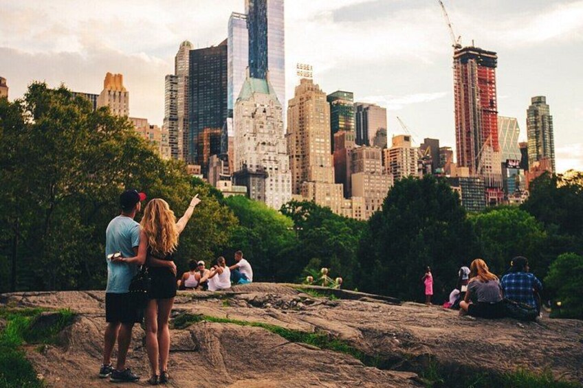 A city skyline with tall buildings, including a distinctive skyscraper with a green spire, and a group of people on a rocky outcrop. 1