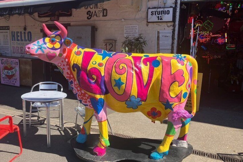 A colorful cow statue with the word 'LOVE' on its side, placed on a street with a chair and a sign in the background. 2
