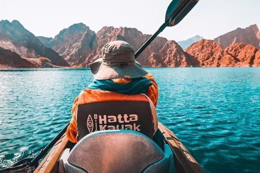 A person kayaking with a hat and scarf, wearing a jacket with 'Hatta Kayak' on the back, surrounded by calm water and mountains. 2