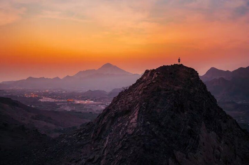 A mountain peak at sunset with a person standing on top, a valley below, and distant mountains. 3