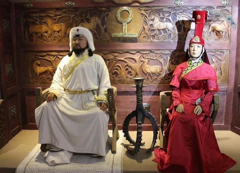 Two individuals seated on ornate chairs in a room with carved wooden panels and a decorative sword. 1