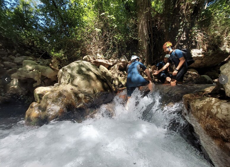 Two people in blue wetsuits wading through a rocky stream surrounded by trees. 3