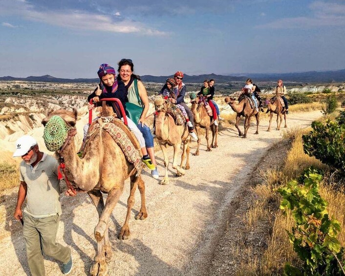 A group of people riding camels on a dirt path with a mountainous landscape in the background. 2