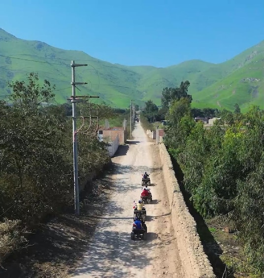 A dirt road flanked by greenery and hills, with a few motorbikes traveling down it. 1