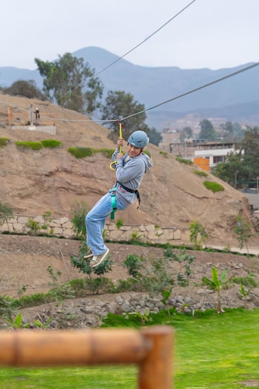 A person ziplining with a harness and helmet, overlooking a hilly landscape with sparse vegetation and a few buildings in the distance. 5