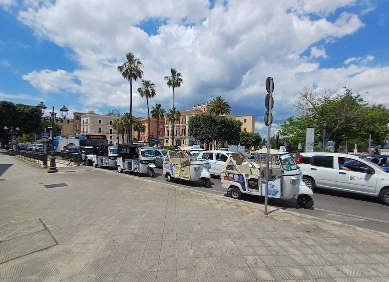 A street with parked tuk-tuks, cars, and a bus. There are palm trees and buildings in the background. 1