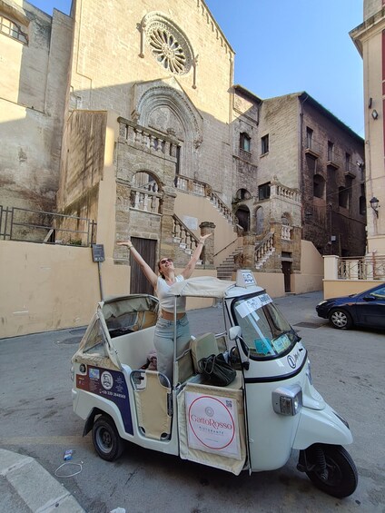 A white three-wheeled vehicle parked in front of a historic building with a large circular window and a staircase leading to an upper level. 2