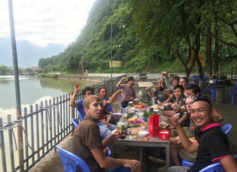 A group of people sitting at a table outdoors by a river, with a forested hill in the background. 3