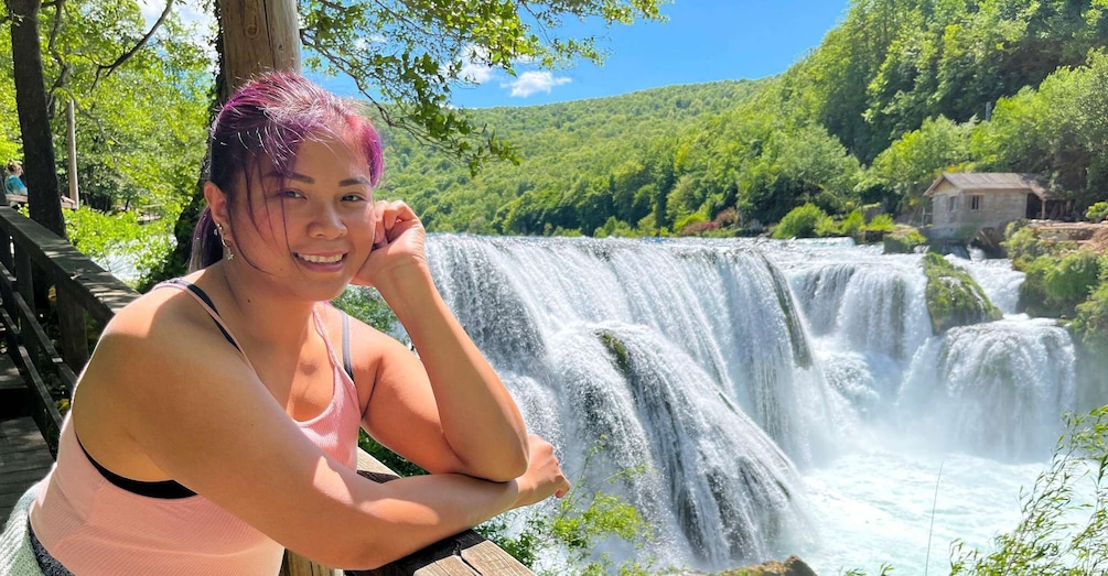 A person with purple hair sitting by a waterfall surrounded by greenery and a small building. 3