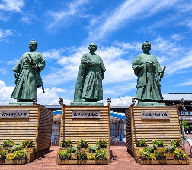 Three statues of historical figures with plaques and potted plants in front. 1