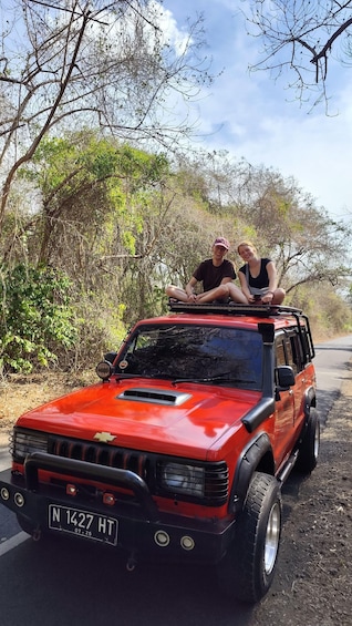 A red off-road vehicle with two people sitting on top, parked on a dirt road surrounded by trees. 1