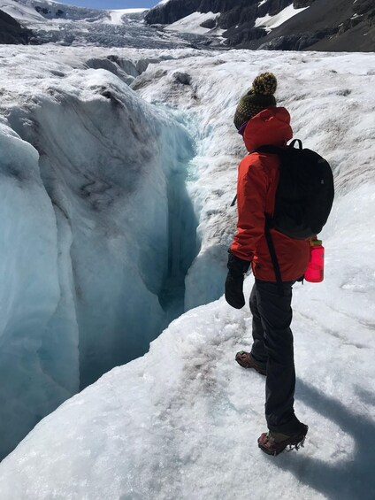 A person in a red jacket and black pants standing on a snow-covered glacier, looking at a blue ice crevasse. 2