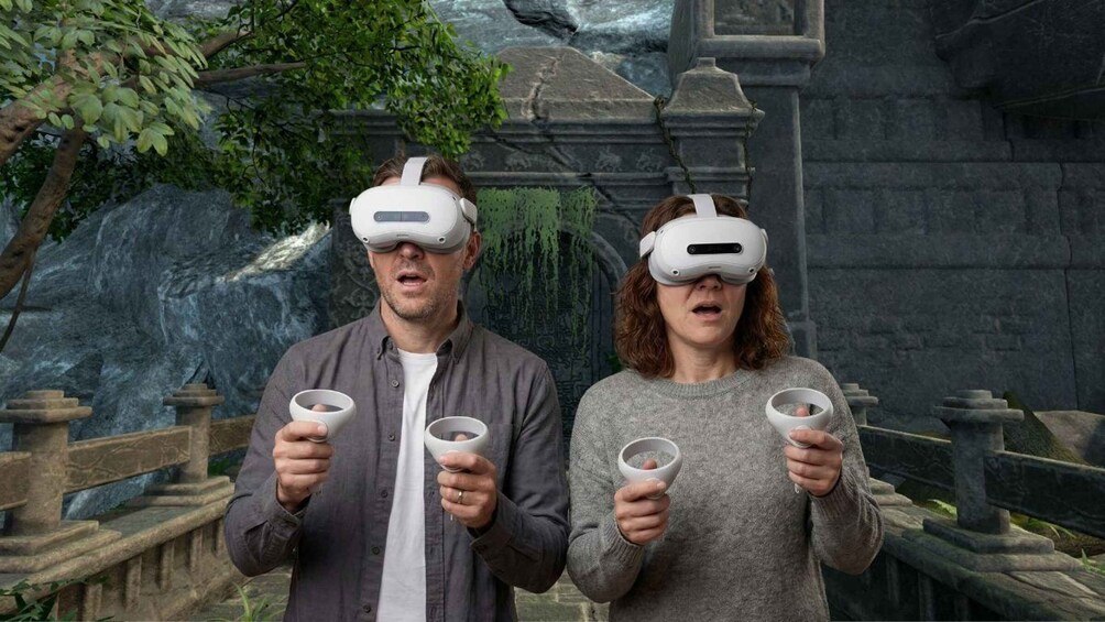 Deux personnes portant des casques de réalité virtuelle, tenant des tasses blanches dans un environnement virtuel composé de structures en pierre et de verdure. 3