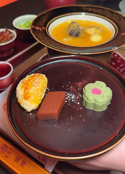 Una mesa con un bol oscuro que contiene un objeto rojo rectangular, un objeto verde con un adorno rosa y un objeto amarillo con semillas de sésamo. 3