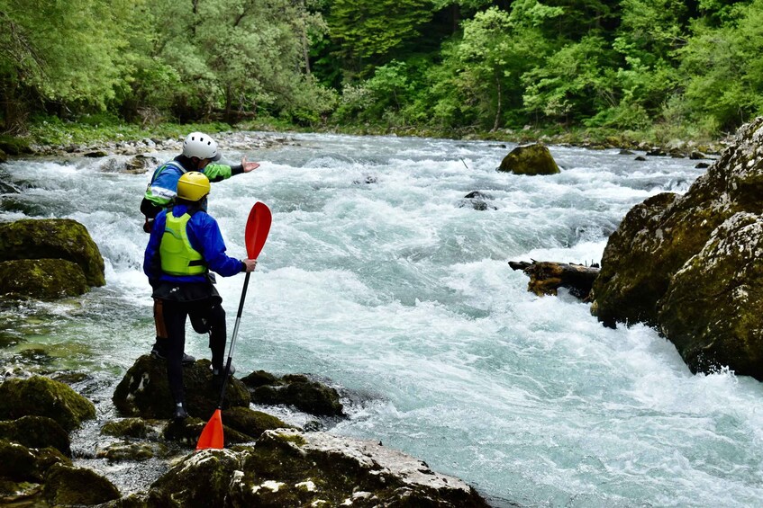 A person in a blue jacket and yellow helmet standing on rocks by a fast-flowing river, holding a red paddle. 3
